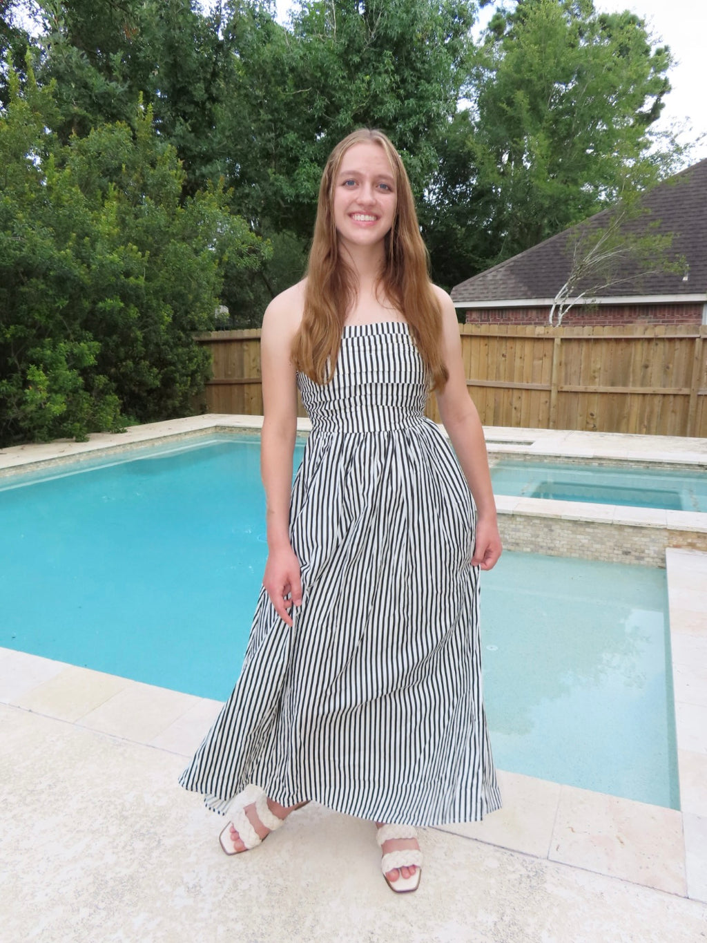 A woman wearing a black and white vertically striped strapless maxi dress stands outdoors next to a swimming pool.