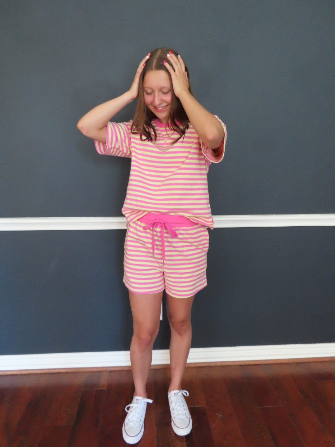 A girl wearing a pink and yellow striped set with a bright pink drawstring, white sneakers, and her hands on her head, standing in front of a dark gray wall.