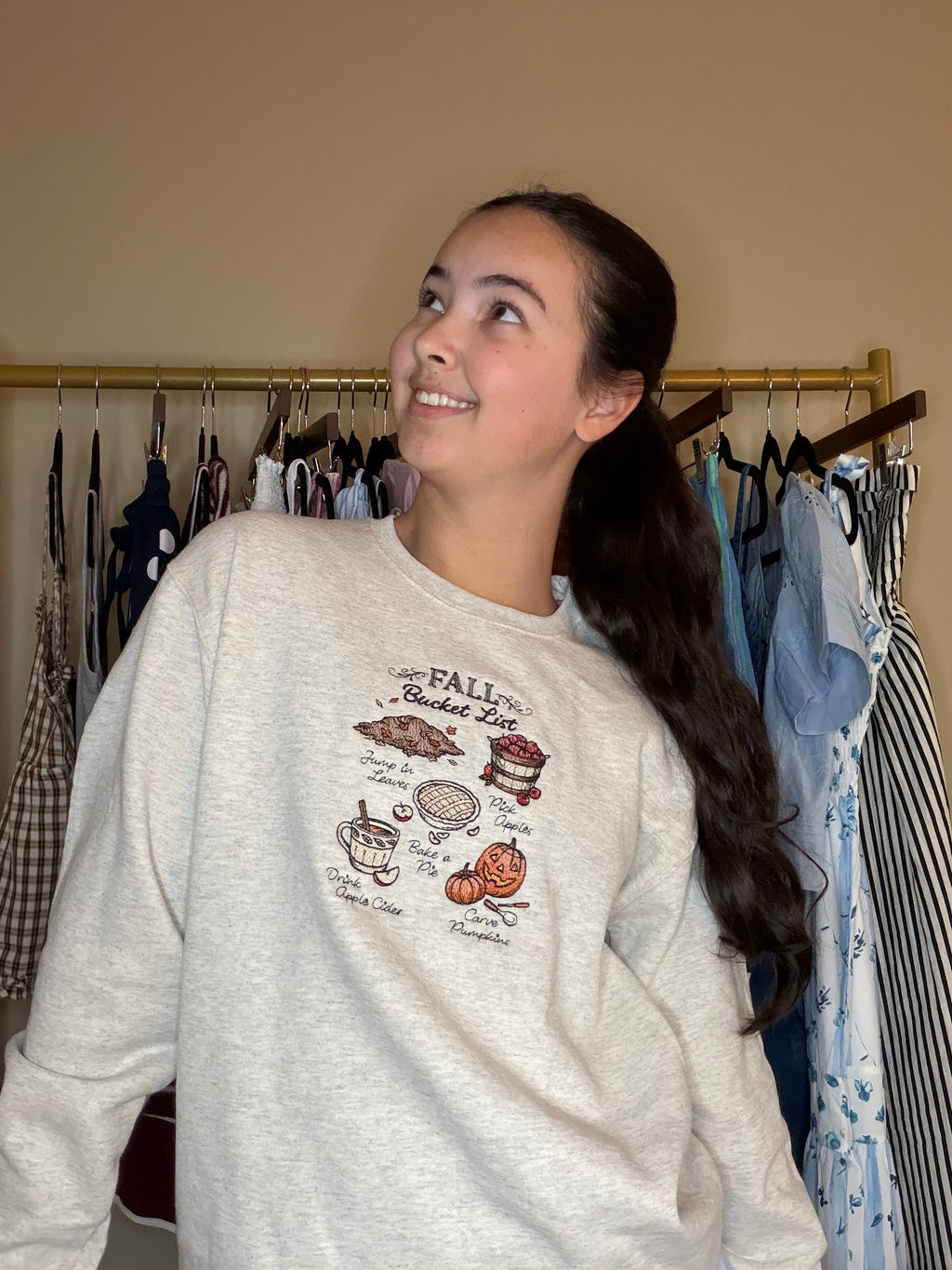 A woman wearing a light gray crewneck sweatshirt with a 'Fall Bucket List' graphic print, standing in front of a clothing rack.  There is embroidered leaves, apples, a pie, apple cider, and pumpkin carving on the sweatshirt.