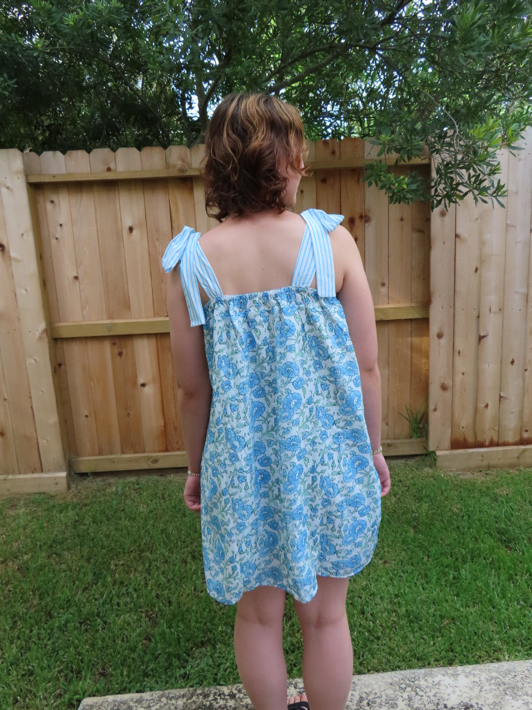 A woman pictured from the back wearing a light blue and white floral print sleeveless dress with tie-shoulder straps, standing outdoors in front of a wooden fence and greenery.