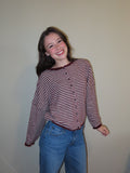 A smiling young woman wearing a long-sleeve button-front top with horizontal burgundy and white stripes, paired with blue jeans, standing against a plain beige wall.