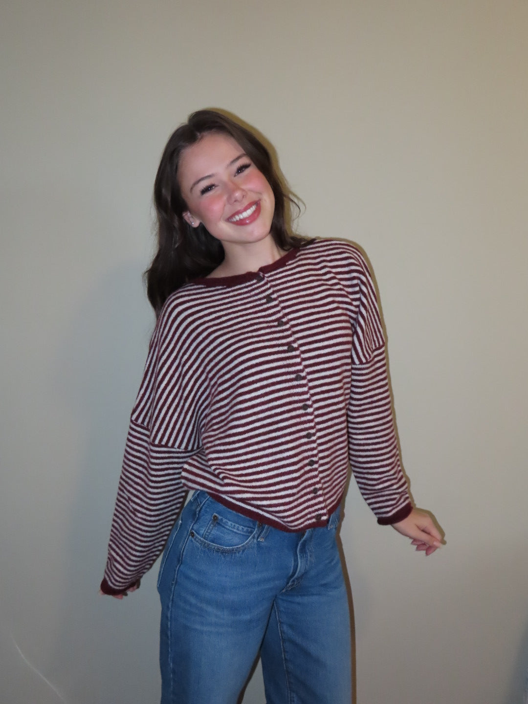 A smiling young woman wearing a long-sleeve button-front top with horizontal burgundy and white stripes, paired with blue jeans, standing against a plain beige wall.