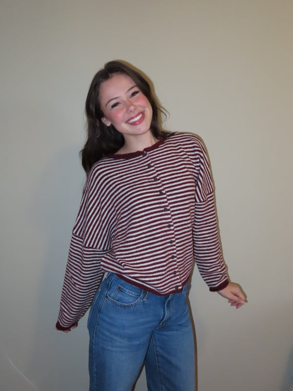 A smiling young woman wearing a long-sleeve button-front top with horizontal burgundy and white stripes, paired with blue jeans, standing against a plain beige wall.