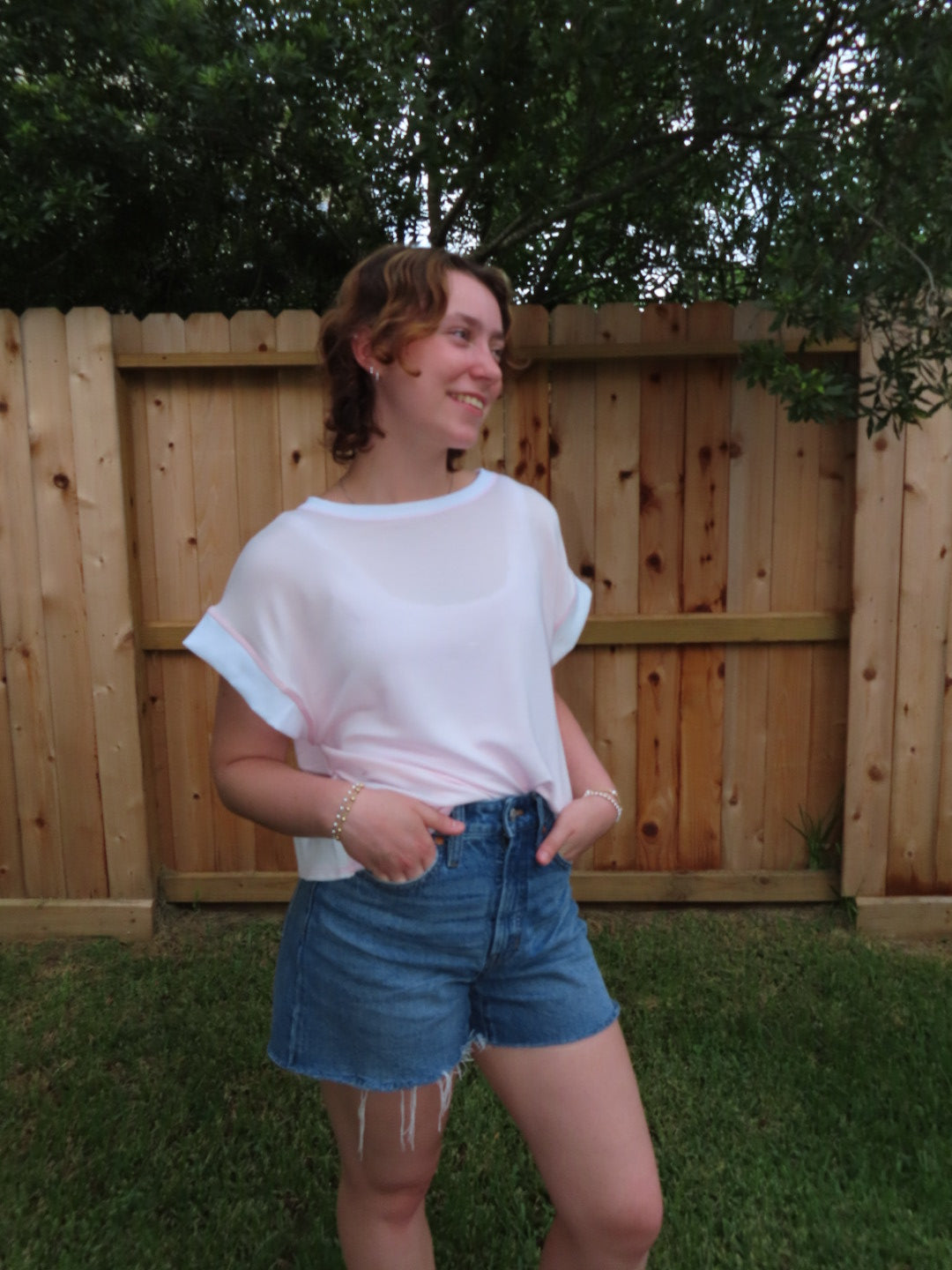 A woman wearing a pink short-sleeve t-shirt and denim shorts, standing outdoors in front of a wooden fence and green grass.