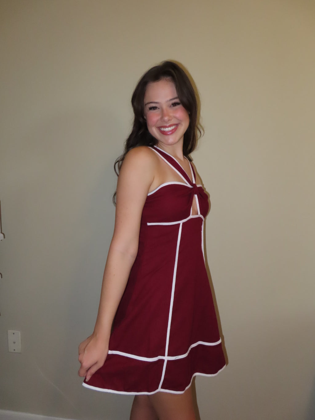 A woman wearing a sleeveless maroon mini dress with white contrast trim, standing against a plain beige wall.