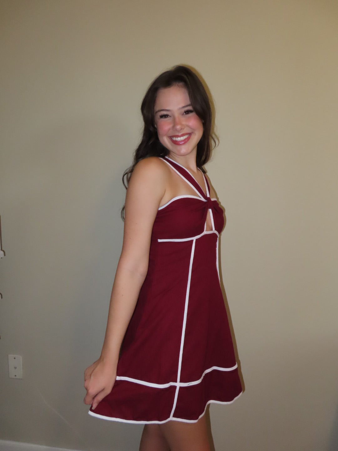 A woman wearing a sleeveless maroon mini dress with white contrast trim, standing against a plain beige wall.
