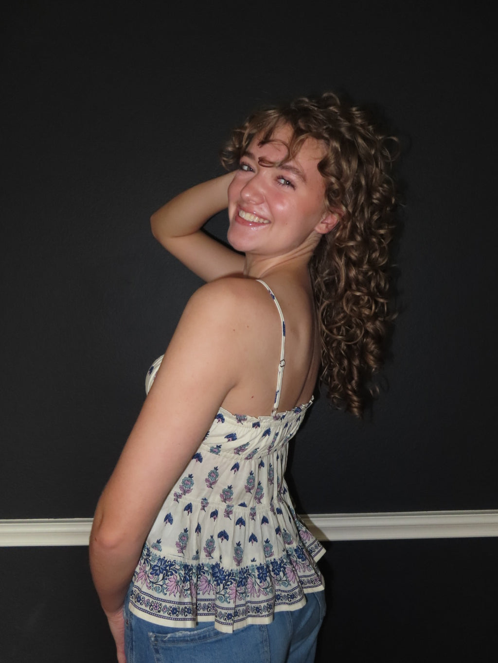A smiling woman with curly hair poses in a creme spaghetti-strap top with a blue and purple floral pattern and blue jeans against a dark background.