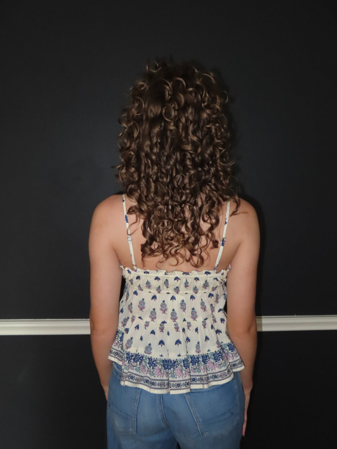 A woman with long, curly brown hair is shown from behind, wearing a creme spaghetti-strap top with a blue and purple bohemian-style floral pattern and blue jeans, standing against a dark gray wall.