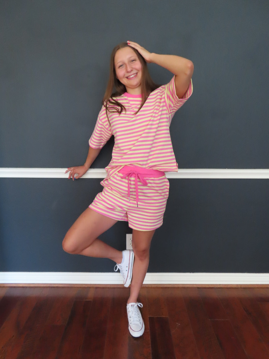 A young girl wearing a pink and yellow striped set and white sneakers, posing in front of a dark gray wall next to a wooden floor.