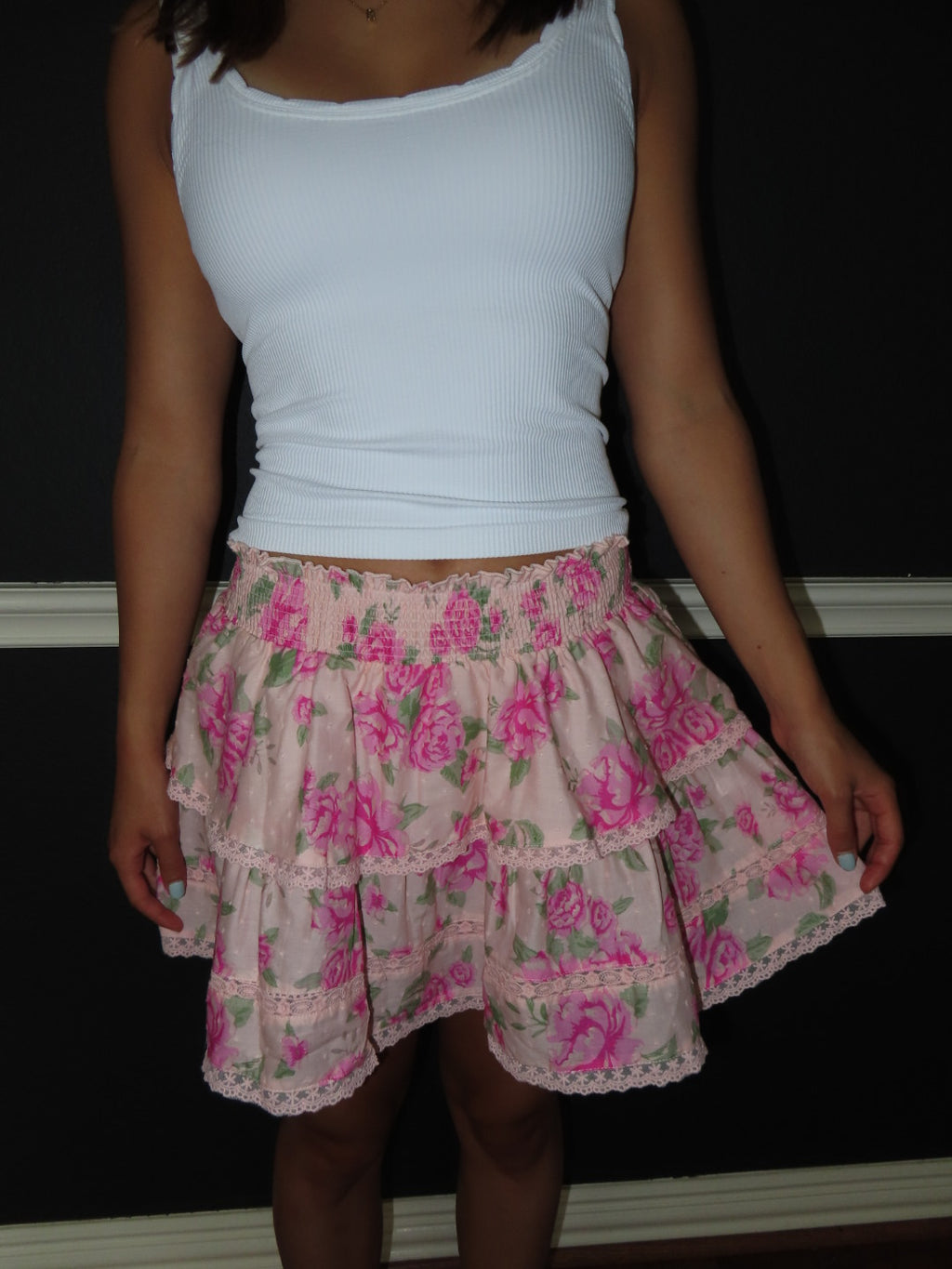 A person wearing a white ribbed tank top and a tiered pink floral skirt with green leaves and lace trim, standing against a dark wall.