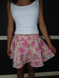 A person wearing a white ribbed tank top and a tiered pink floral skirt with green leaves and lace trim, standing against a dark wall.