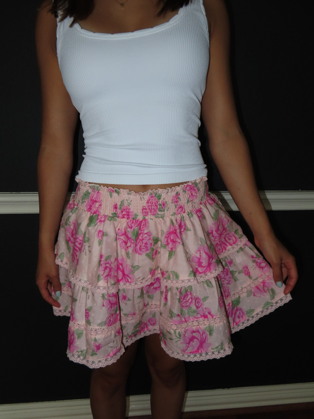 A person wearing a white ribbed tank top and a tiered pink floral skirt with green leaves and lace trim, standing against a dark wall.