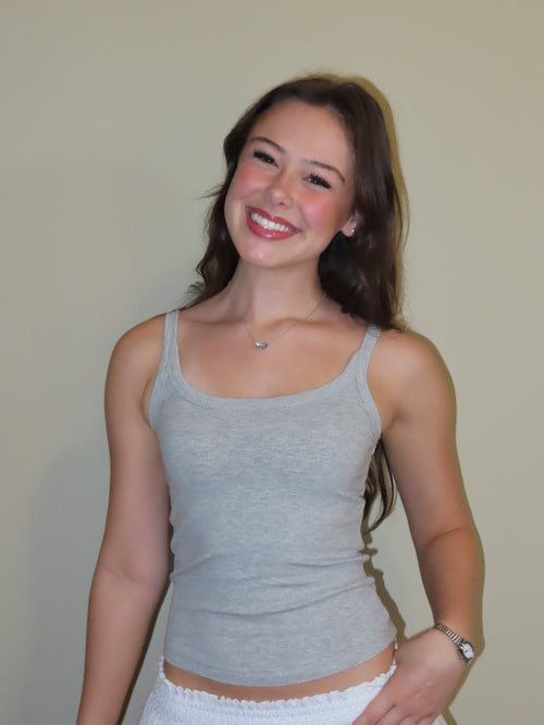 A smiling young woman with long brown hair wearing a light gray ribbed tank top and white bottoms, standing against a plain beige wall.