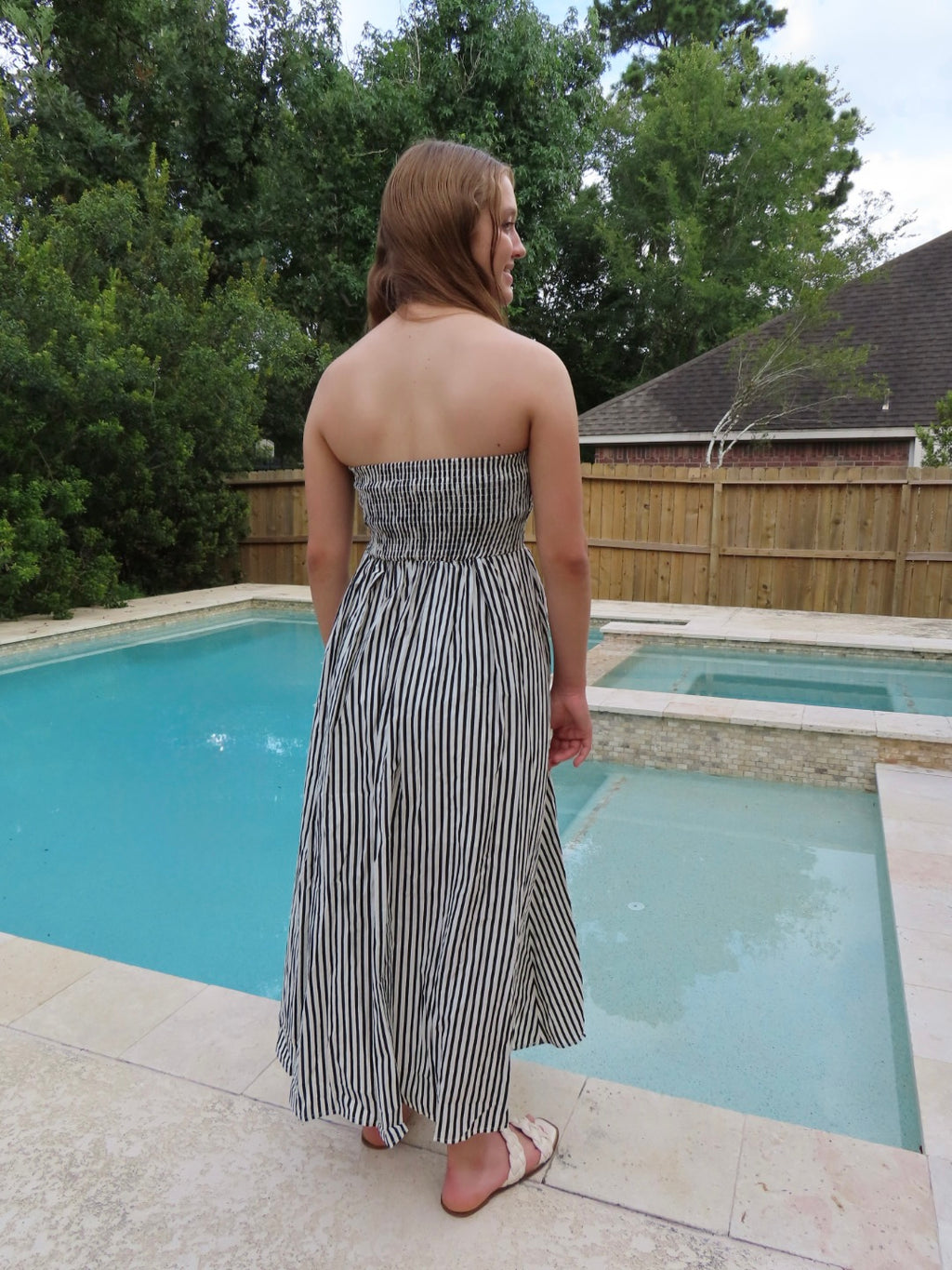 A woman stands with her back to the camera by a swimming pool, wearing a strapless black and white vertically striped maxi dress.